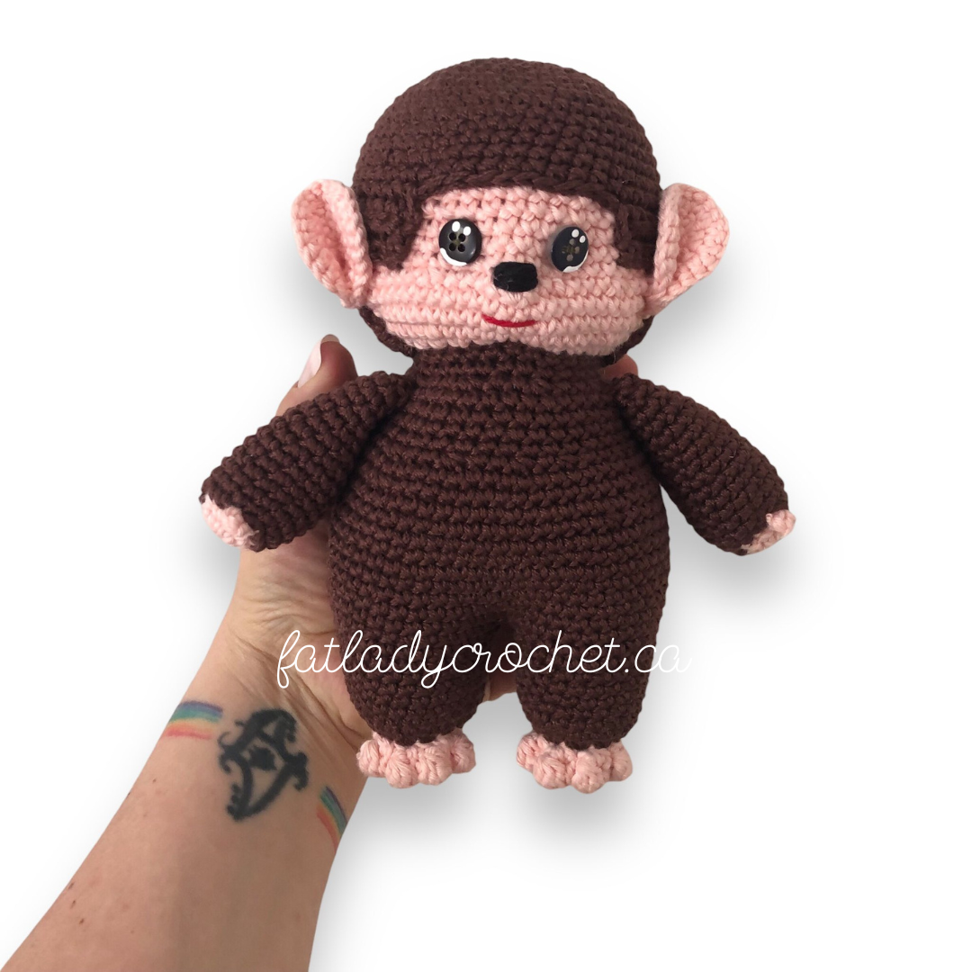 A crocheted monchhichi doll behind held up against a white background. 