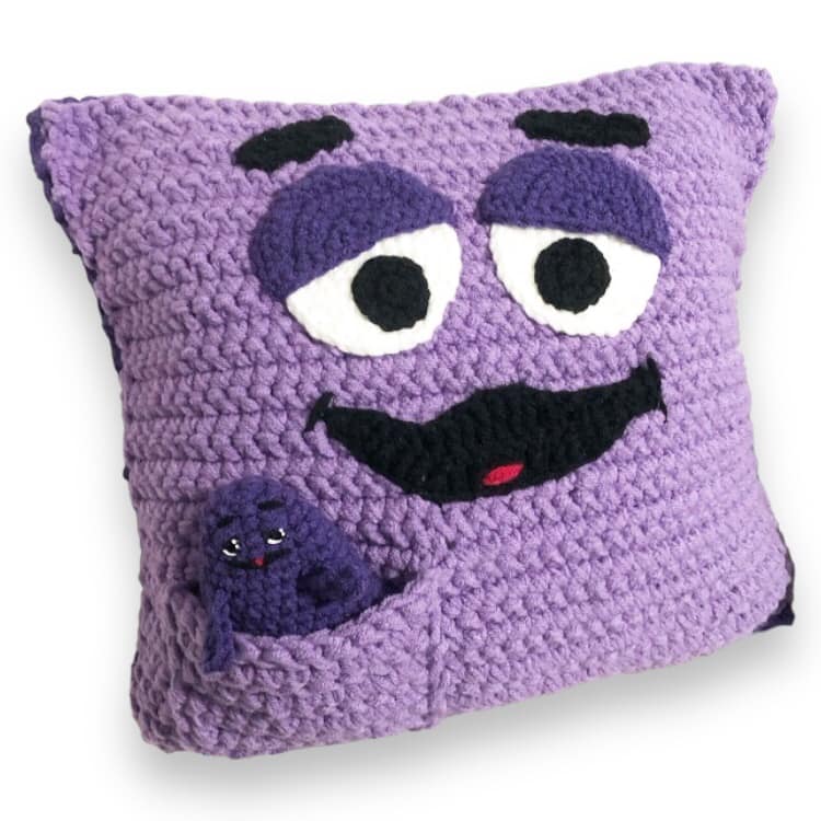 A crocheted doll and pillow both resembling Grimace, with the doll of Grimace inside a pocket. 