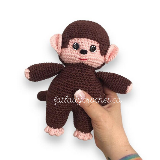 A crocheted monchhichi doll being held by a hand against a white background with Fat Lady Crochet branding.