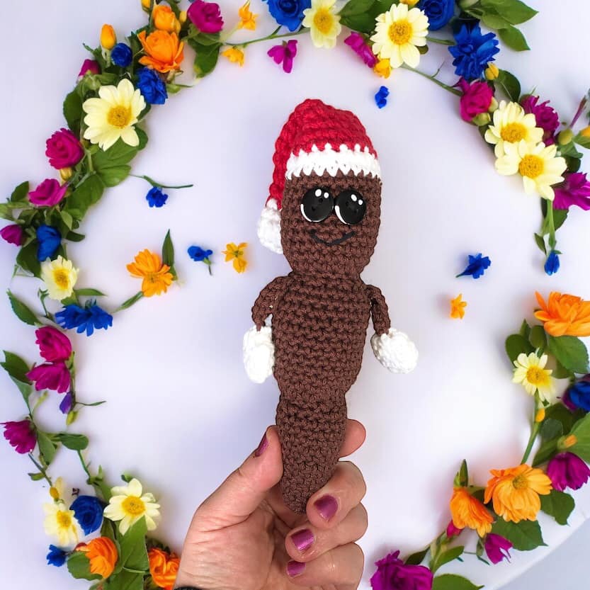 A hand holding up a crocheted doll of Mr. Hanky the Christmas Poo against a background with a flower wreath.
