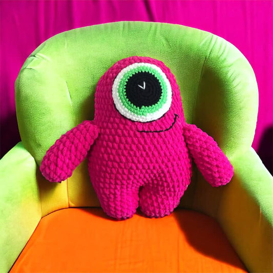 A pink crocheted monster plushie with one green eye sitting on a green and orange chair. 