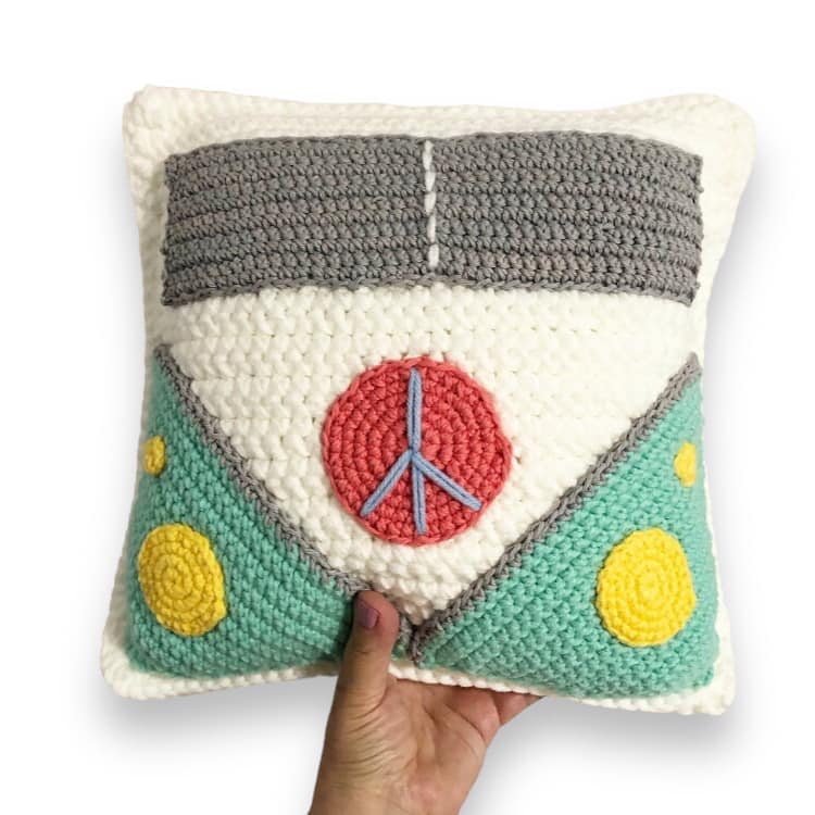 A crochet cushion with a van design, featuring peace symbols and multiple colors including white, grey, turquoise, coral, and yellow being held up against a white background. 