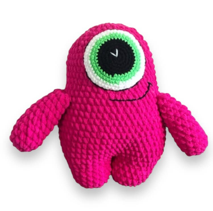 A pink crocheted monster plushie with one green eye standing against a white backround. 