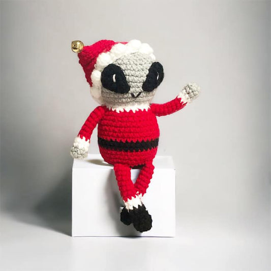 A crochet grey alien dressed like Santa Claus sitting on a cube and waving. 