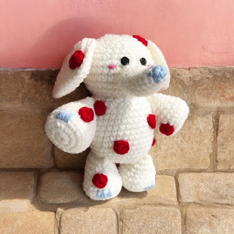 A white and red spotted crochet elephant from Rudolph the Red Nosed Reindeer standing against a pink and beige brick wall. 