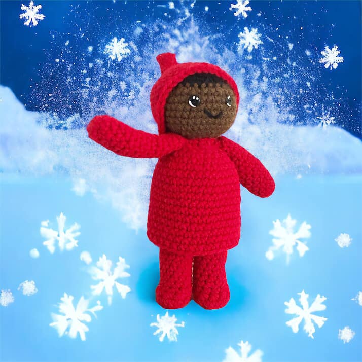 A crocheted doll resembling Peter from the Snowy Day by Ezra Jack Keats standing in the snow surrounded by snowflakes. 
