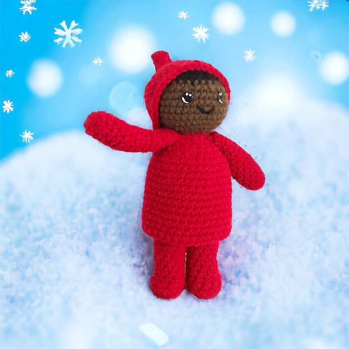 A crocheted doll resembling Peter from the Snowy Day by Ezra Jack Keats standing on a snowy bank surrounded by snowflakes. 
