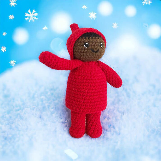 A crocheted doll resembling Peter from the Snowy Day by Ezra Jack Keats standing on a snowy bank surrounded by snowflakes. 