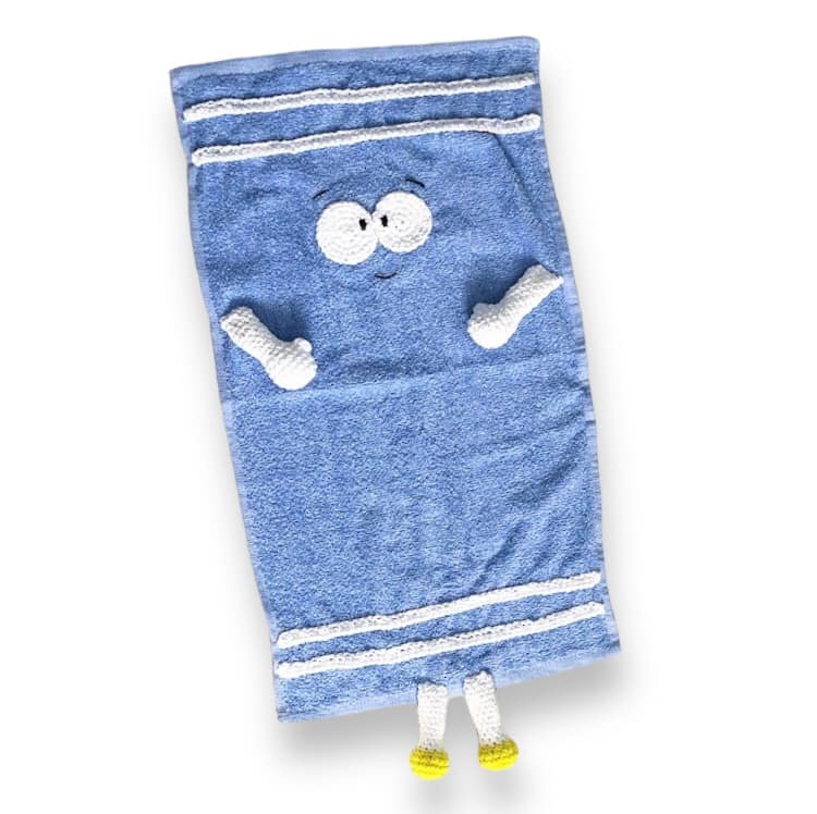 A hand towel with crocheted add-ons made to resemble Towelie from South Park against a white background. 